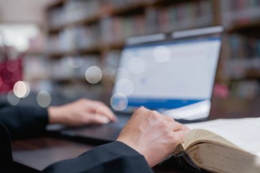Sessiz bir masada, açık bir dizüstü bilgisayar ve kitapla yoğun bir çalışma oturumu yürütüldü, elleri, kütüphane içinde bir makaleyi eşelemeye hazır.
