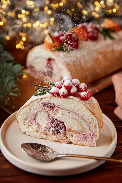 Slice of festive meringue roll with cherries decorated for Christmas with berries, citrus and rosemary on wooden table.