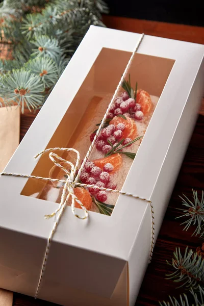 Festive Christmas meringue roll with cherries decorated with berries, citrus and rosemary packed as present or food delivery on wooden table.