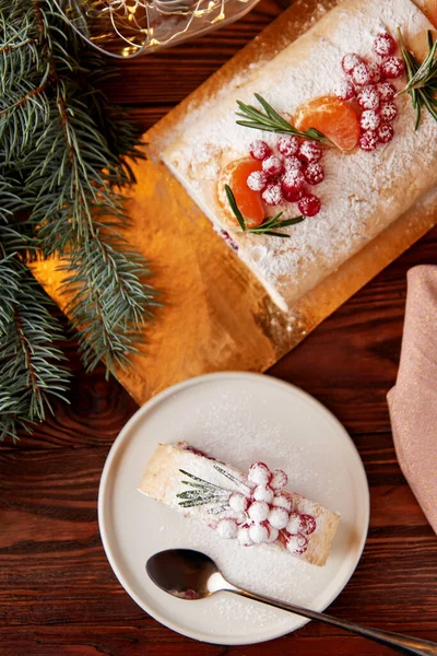 Slice of festive meringue roll with cherries decorated for Christmas with berries, citrus and rosemary on wooden table.