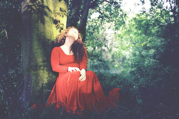 Lady with red dress falls asleep angelically leaning against a tree in a forest