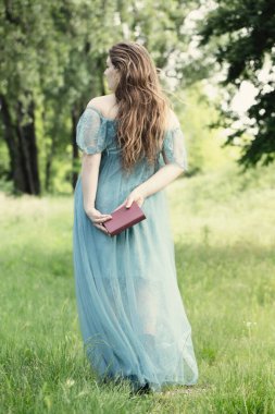 Rönesans tarzında zarif bir kadın elinde kitapla çayırda yürüyor.