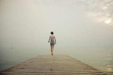 Young girl from behind walking on the pier towards the sea, concept of freedom and travel