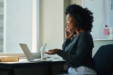 Dost canlısı Afrikalı Amerikalı iş kadını müşterilerle telefonda konuşuyor, dizüstü bilgisayar kullanıyor.