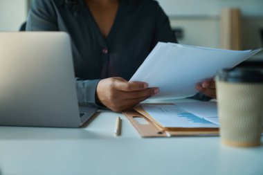 Çok ırklı bir kadın iş yerinde dizüstü bilgisayarın önünde oturuyor ve elinde çalışma kağıtlarını tutuyor.