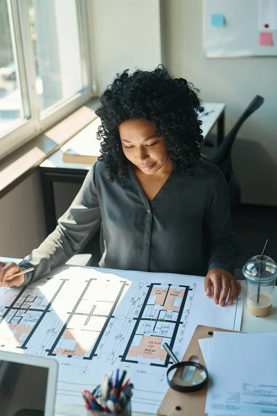 絹のブラウスで穏やかなアフリカ系アメリカ人の女性は設計図と大きな窓の近くのオフィスデスクで動作します — ストック写真