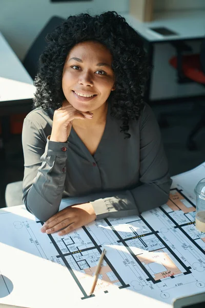 Multiracial female designer in a gray blouse sits at her desk and looks at the camera with a pleasant smile