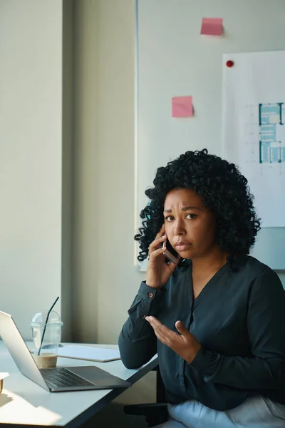 Worried multiracial woman in gray blouse sitting at workplace and talking on phone