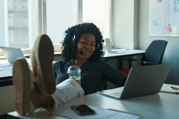 モバイルヘッドセットで多人種コールセンターの演算子を笑顔テーブルの上に彼女の足を置き 積極的に発信者と通信します — ストック写真