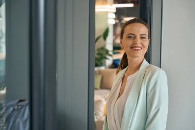 Birlikte çalışma alanında duran ve kameraya neşeyle bakan zarif genç iş kadınının portresi.