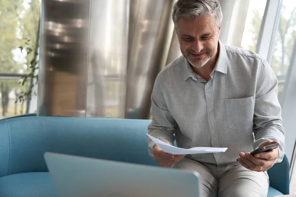 Middle-aged company manager reads business papers with a satisfied look, he is located in a comfortable coworking