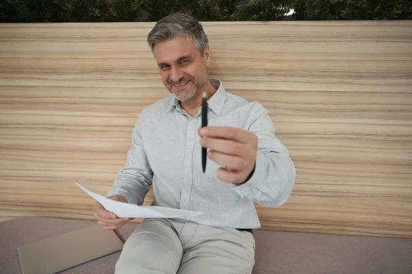 Smiling manager extends a pen to a subordinate, he sits against the background of a wooden wall