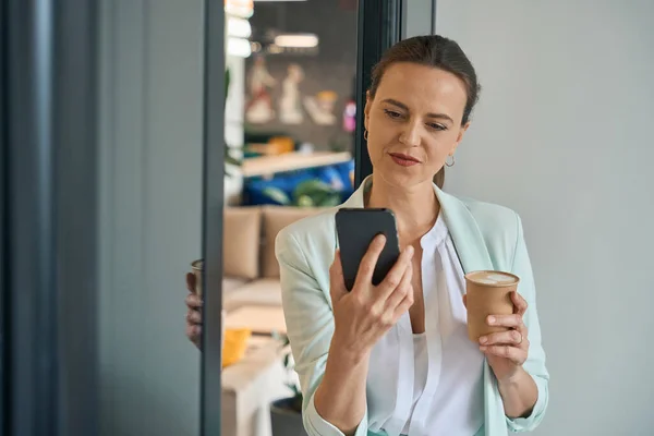 Cheerful elegant lady is standing in office and holding cup of drink in one hand and cell phone in other