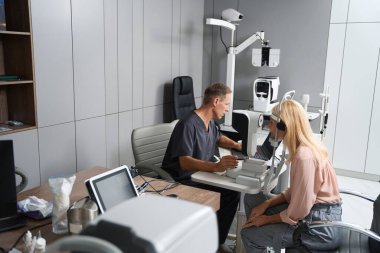 Sandalyede oturan ve özel tıbbi cihazlara bakan bir kadın, karşıda oturan ve sonuçlara bakan bir sağlık görevlisi.