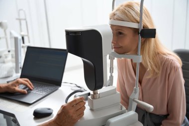 Kadın sandalyede oturuyor ve özel tıbbi cihazlara bakıyor. Doktor karşıda oturuyor ve bilgisayarla uğraşıyor.