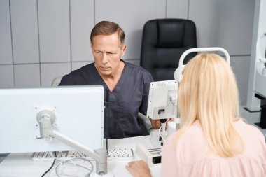 Hasta sandalyeye oturuyor ve özel tıbbi cihazlara bakıyor. Doktor karşıda oturuyor ve bilgisayara bakıyor.