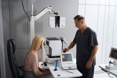 Kadın sandalyede oturuyor ve özel tıbbi cihaza bakıyor, doktor ekranda dikiliyor ve bakıyor.