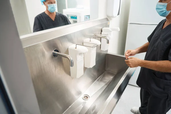 Doctor in black medical suit and protective mask standing near sink with disinfectants and prepare to wash hands