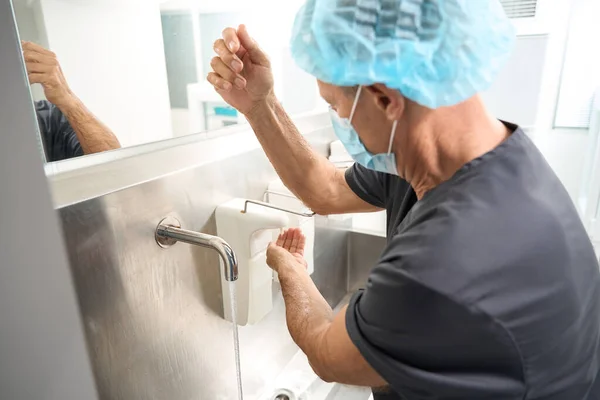 Male doctor in black medical suit and protective mask standing near sink with disinfectants and washing hands
