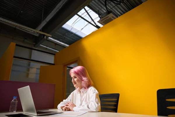 有一头粉色头发的年轻女性坐在办公室的桌子旁边 看着笔记本电脑 黄色明亮的背景 — 图库照片
