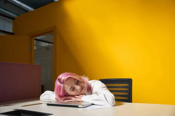 有一头粉色头发的女士坐在办公室的桌子旁边 躺在笔记本电脑上 黄色明亮的背景 — 图库照片