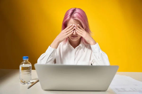 一个有着粉色头发的悲哀女士坐在桌子旁边 办公室里开着笔记本电脑 背景是黄色的 — 图库照片