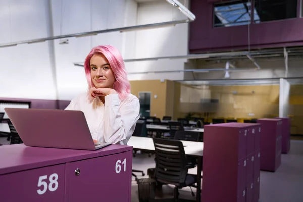 粉红头发的设计师坐在桌旁 手握手提电脑 手牵手地看着摄像机 — 图库照片