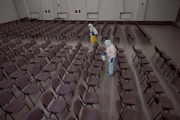 Two professional cleaners in overall protective garments spraying chemicals for disinfection on chairs in conference room
