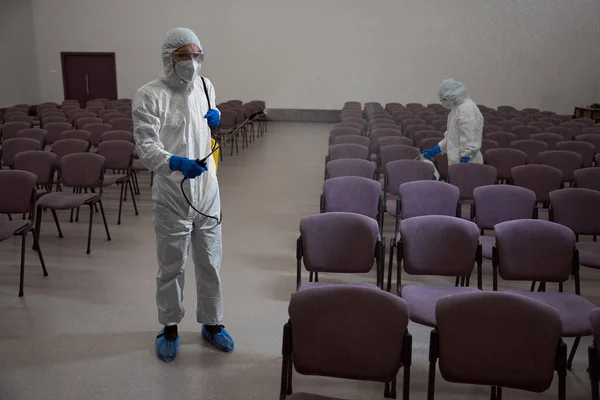 Certified cleaners in protective garments and masks spraying disinfectant liquid over the chairs in the building