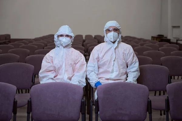Two people in full-body protective suits, masks and goggles sitting together on chairs and looking at the camera