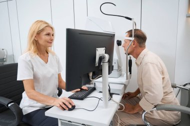 Beyaz önlüklü profesyonel bir doktor bilgisayarın başında oturuyor ve hastaların göz muayenesini hastanede yapıyor.