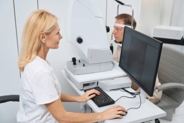 Beyaz önlüklü gülümseyen hemşire bilgisayarın başında oturuyor ve dolapta göz muayenesi yapıyor.