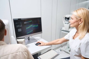 Bilgisayarın başında oturan beyaz önlüklü doktor hastaya göz küresi resimleri gösteriyor.