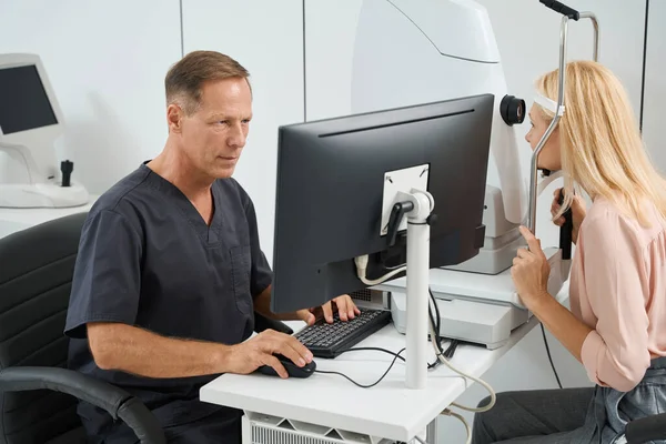 Doctor in black coat sitting at the computer and performing eye examination of eyeball for lady in the hospital