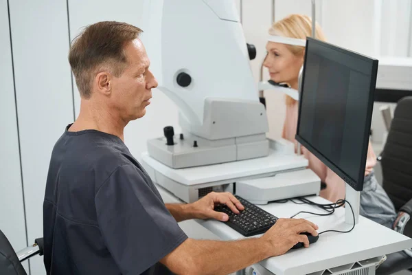 Ophthalmologist in black coat sitting at the computer and performing eye examination of eyeball for lady in the hospital