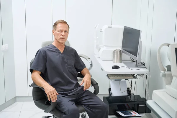 Tıbbi Kıyafetli Bir Erkek Doktor Masada Bilgisayarla Oturuyor Kameraya Bakıyor — Stok fotoğraf