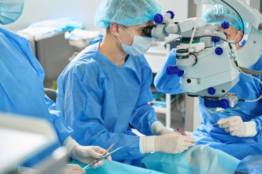 Koruyucu kıyafetli, maskeli ve şapkalı genç doktor dürbünle cihazın yanında duruyor ve hastaya bakıyor.