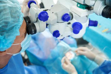Korumacı, maskeli ve şapkalı bir tıp çalışanı cerrahi mikroskoba bakıyor ve hastaya bakıyor.