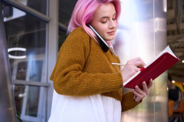 Ofiste duran pembe saçlı genç bir kadının portresi. Elinde defter, kalem ve yazı var.