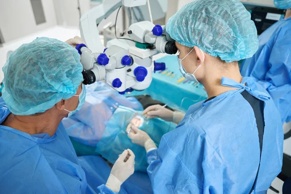 Professional doctor in protective , mask and hat looking in surgical microscope on patient