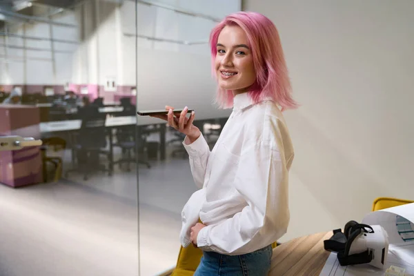 stock image Pretty woman with pink hair holding phone, speaking with client and looking at the camera in coworking