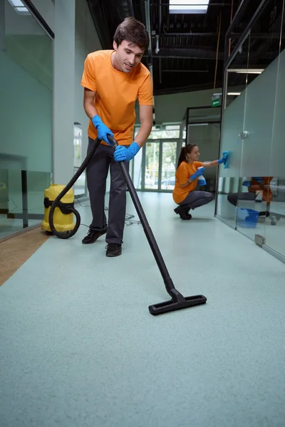 stock image Enthusiastic janitor using the vacuum-cleaner on the floor while his partner wiping the glass surface