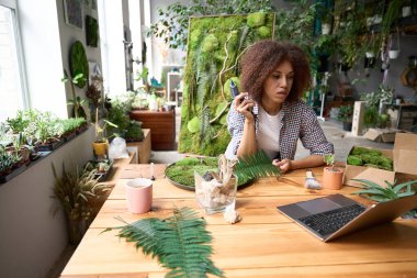 Genç bir kadın masa başında oturmuş dizüstü bilgisayara bakarken yaprakların bileşimini yapmak için aletler kullanıyor.