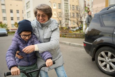 Kışlık elbiseli bir çocuk evin yanında scooter kullanıyor, büyükannesi yanında duruyor ve ona yardım ediyor.