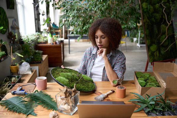 Woman is sitting at desk with notebook and decorative plants and holding moss for making green arrangments