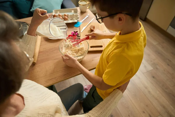 小孩站在厨房的桌子旁边 用面粉打鸡蛋 奶奶坐在旁边加牛奶 — 图库照片