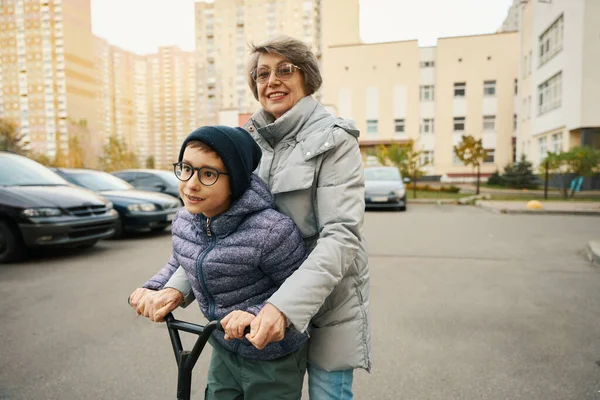 冬の服の子供は 家の近くに祖母とスクーターに乗って楽しんでいます — ストック写真