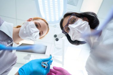 Parlak bir odada genç bir asistan kadın bir cerraha yardım ediyor. Şırınga ve koruyucu maskeler kullanıyorlar.