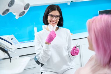 Dost canlısı bir dişçi, dişçi muayenehanesinde bir hastayla iletişim kuruyor. Elinde özel bir alet tutuyor.