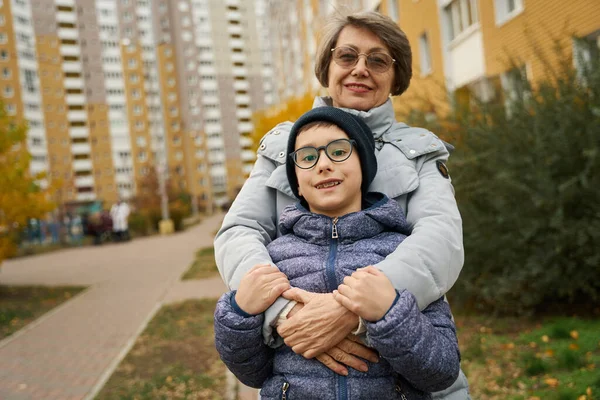 Portrait of happy granny in winter clothes standing and hugging boy outside